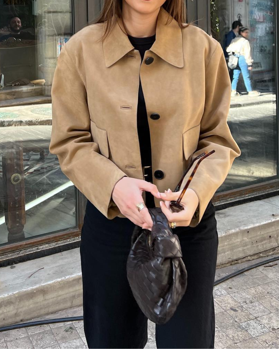 Person wearing a beige jacket holding a brown handbag on a city street.