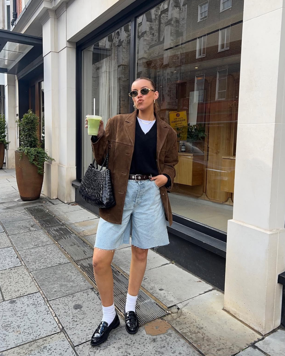 Woman in stylish outfit standing on a city street, holding a drink.