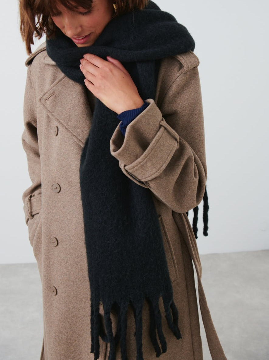 Person wearing a long brown coat and a black scarf with fringes on a plain background
