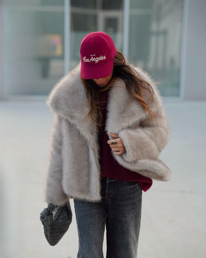 Person wearing a fluffy beige coat, maroon sweater, gray jeans, and pink 'Los Angeles' cap.