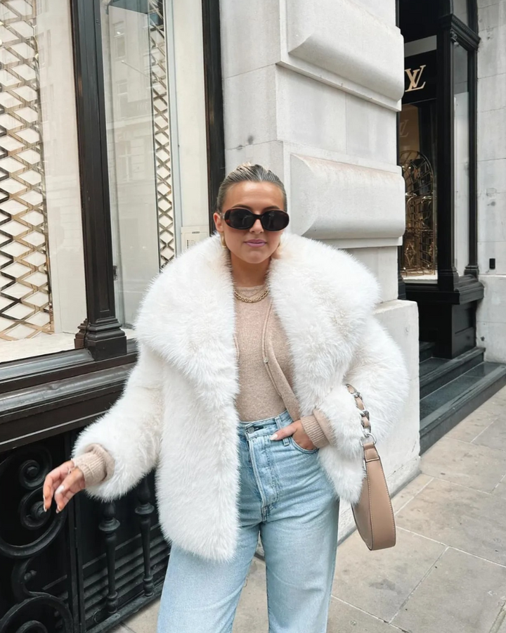 Person wearing a white fur coat, sunglasses, and light blue jeans in front of a building.