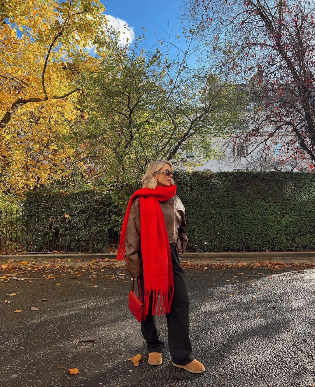 Person wearing a red scarf and beige coat standing on a street with autumn trees in the background