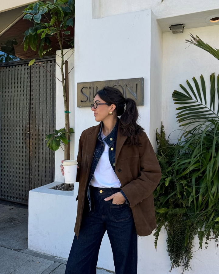 Woman standing outside a building with plants, wearing a brown coat and holding a coffee cup.