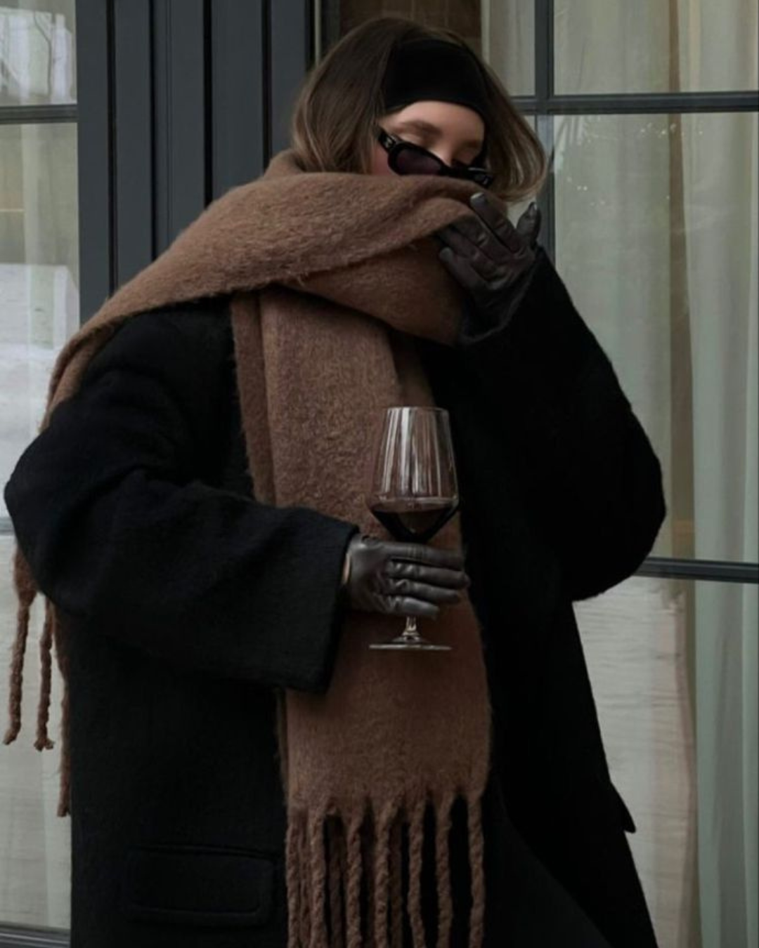 Person wearing a large brown scarf and holding a glass of red wine, standing indoors.
