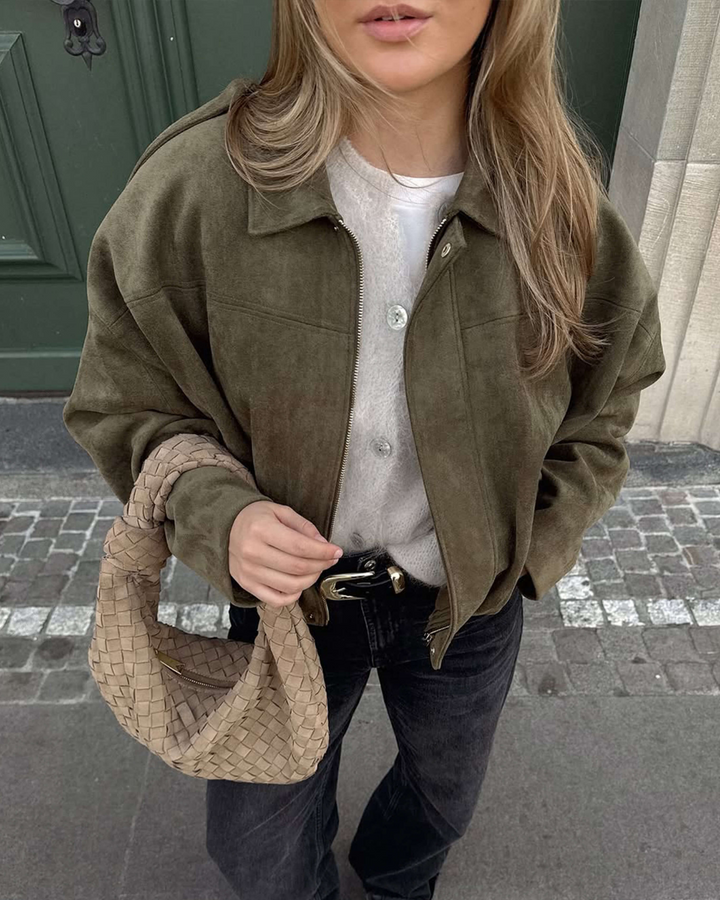 Person wearing a green jacket and holding a beige handbag on a street.