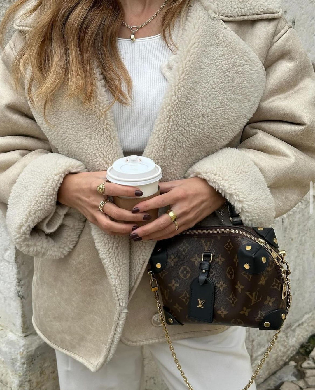 Person wearing a beige coat holding a coffee cup with a Louis Vuitton bag.