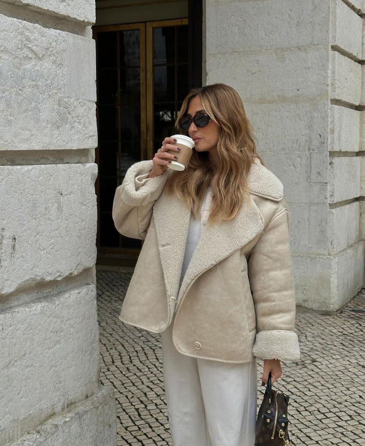 Woman in a beige shearling coat drinking coffee against a stone building.