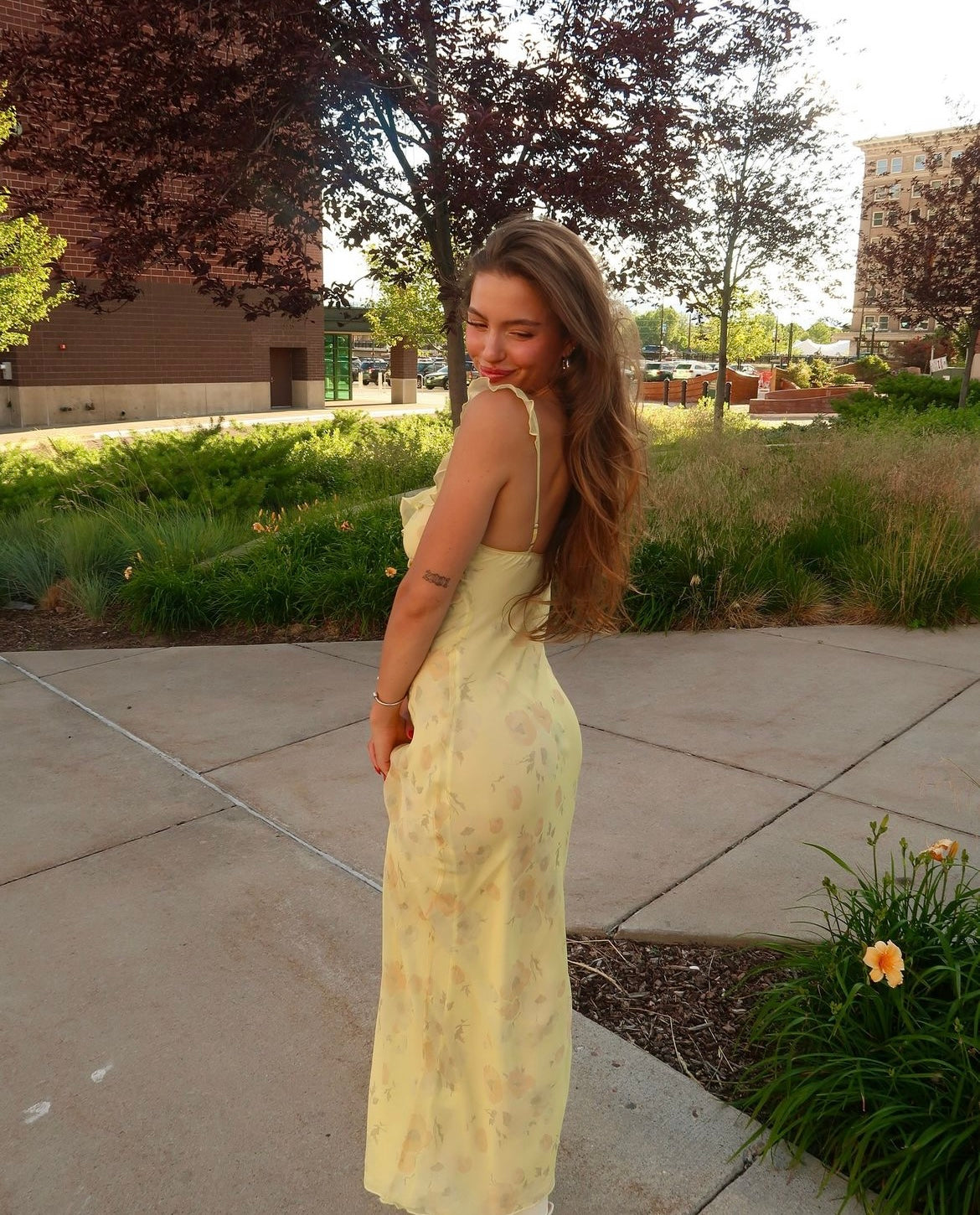 Woman in a yellow dress standing outdoors on a sidewalk with trees and grass in the background