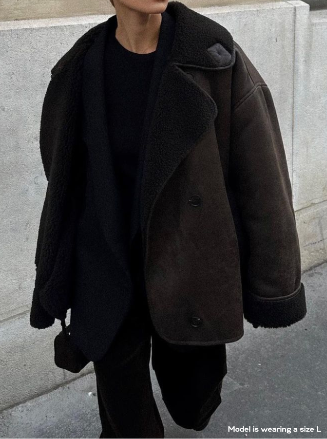 Person wearing a dark brown shearling coat against a concrete wall.