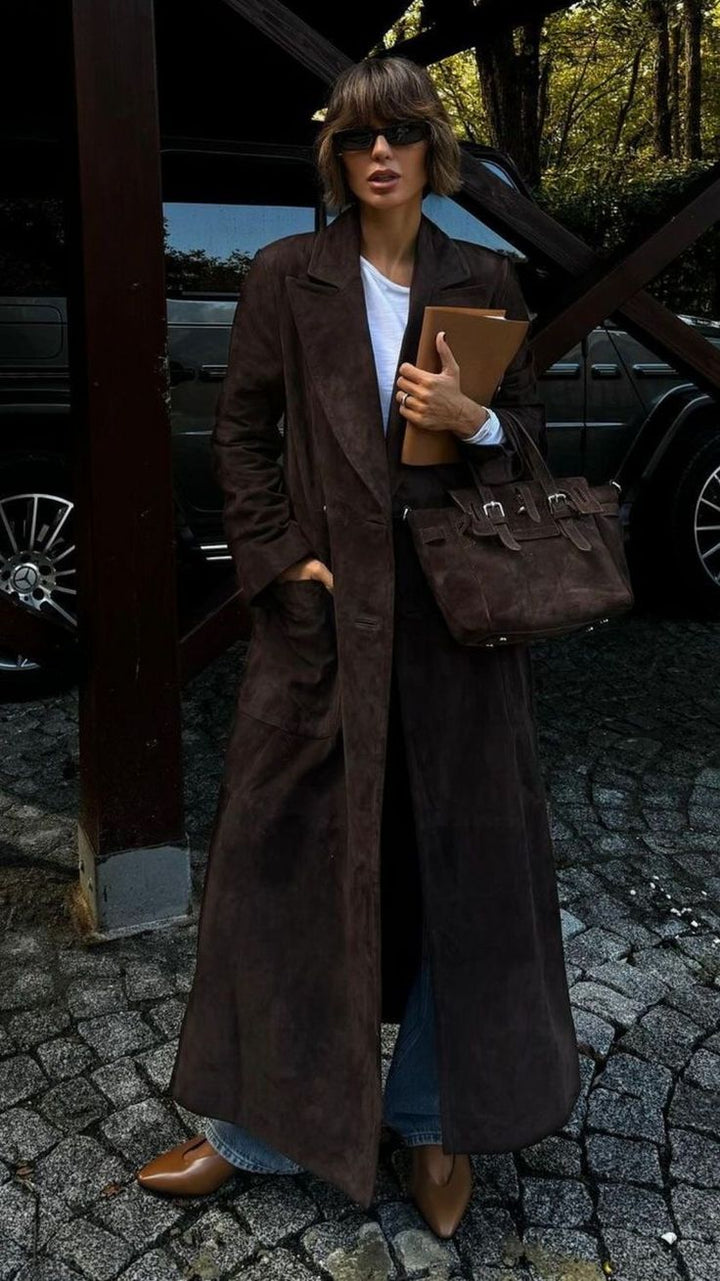 Person wearing a long brown coat holding a brown bag and a folder, standing on a cobblestone street.