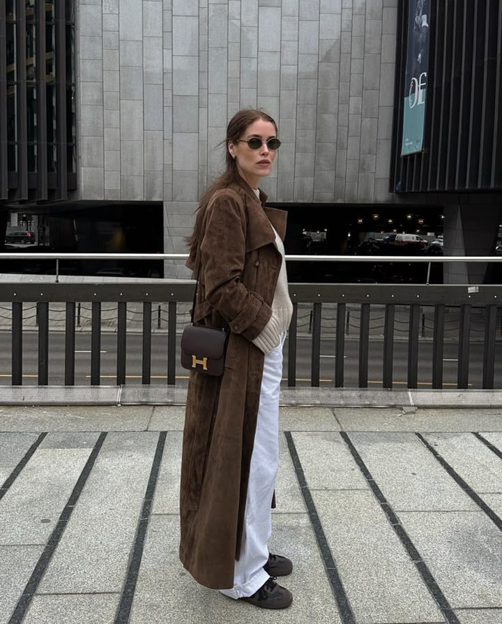 Woman wearing a brown coat and sunglasses, standing on a city street.