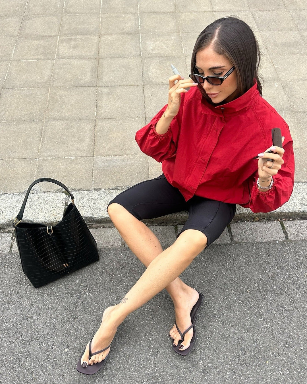 Woman in red shirt and black shorts sitting on a curb with a black handbag next to her.
