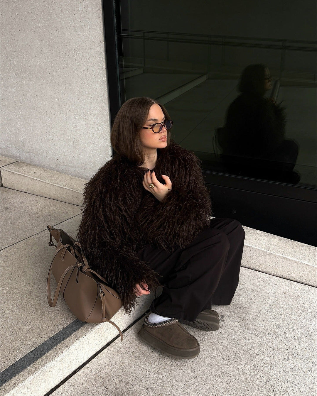 Person sitting on a step wearing a dark fur coat and glasses, with a reflective surface behind them.
